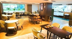 Un comedor con mesas y sillas redondas, una cocina al fondo y vistas a árboles a través de amplios ventanales.