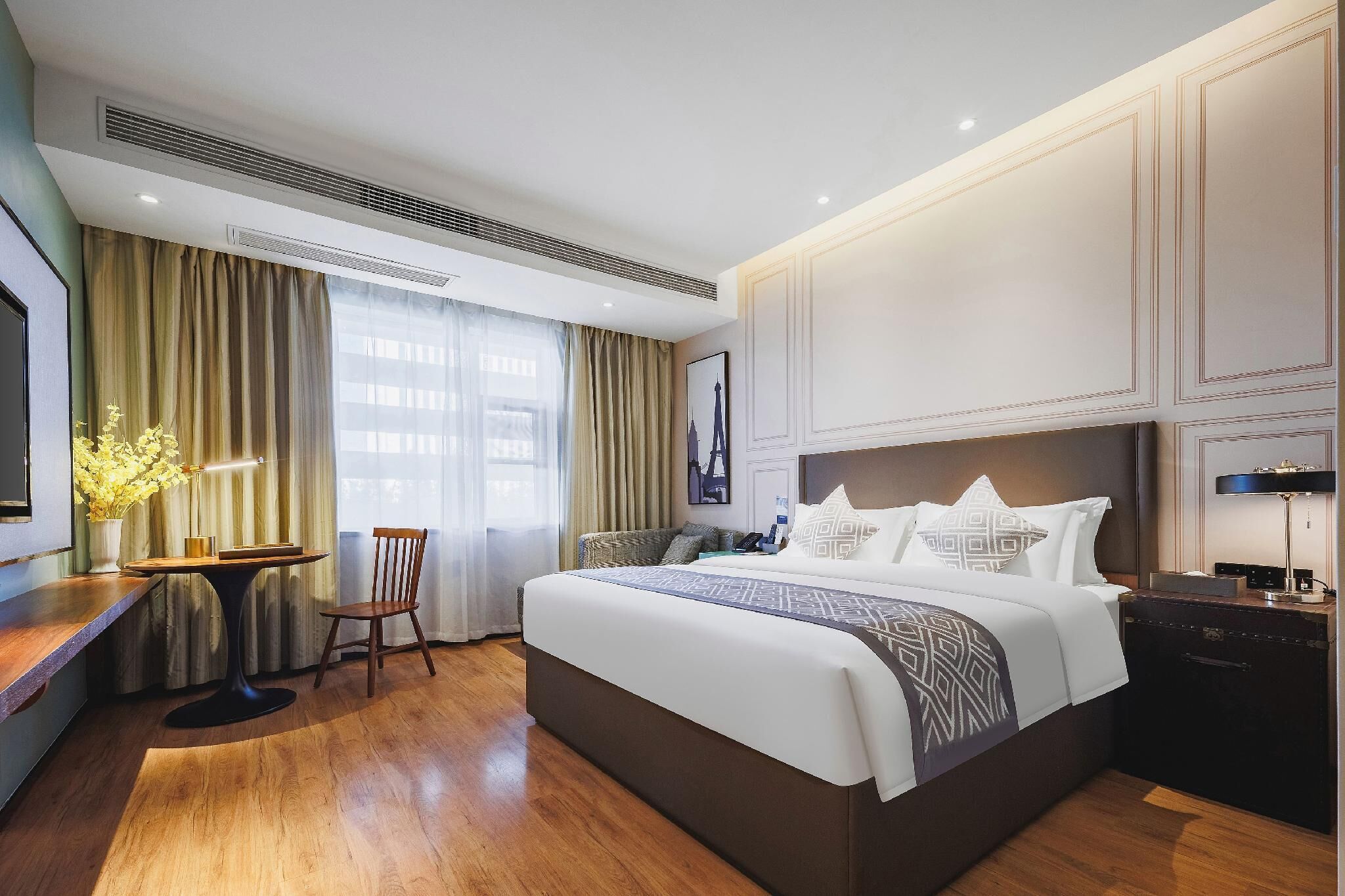 Quarto de hotel com uma cama grande, uma escrivaninha com luminária, uma cadeira e uma janela com cortinas.