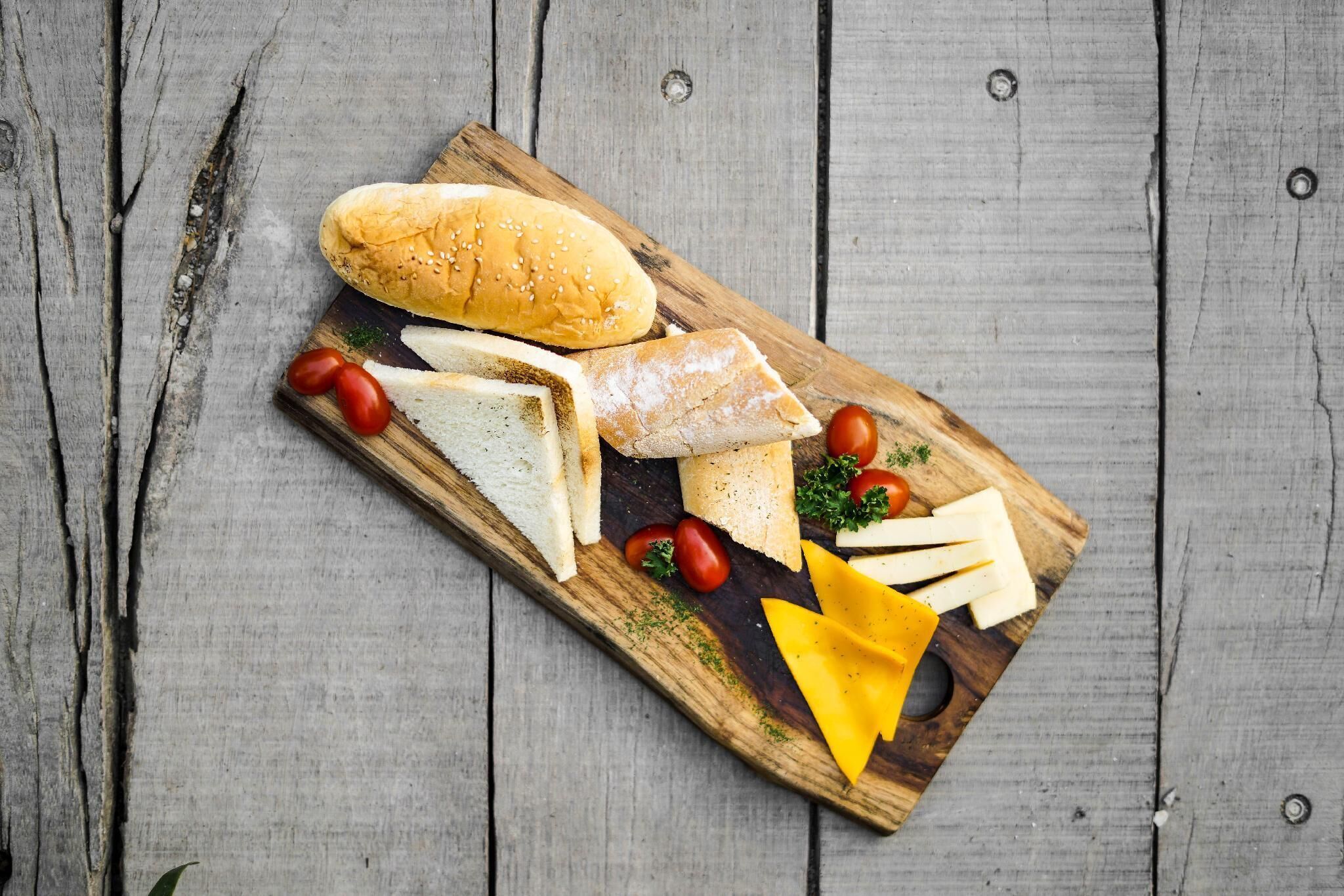 A wooden board with various cheeses, tomatoes, and a baguette on a rustic wooden surface.