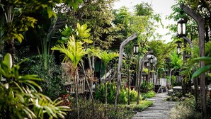 A stone pathway leading through a lush garden with various plants and a traditional wooden structure in the background. - La Luna Resort Yogyakarta (Ngaglik)