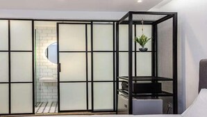 Een kamer met een schuifglazen deur naar een badkamer, een grijs bekleed hoofdeinde en een kleine potplant.