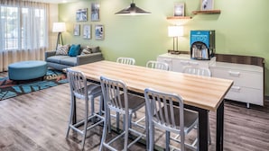 Una habitación de hotel moderna con una mesa de comedor de madera, sillas blancas, un sofá con cojines estampados, una lámpara de pie y una máquina de café.