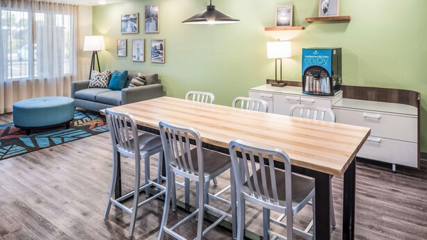 A modern hotel room with a wooden dining table, white chairs, a sofa with patterned cushions, a standing lamp, and a coffee machine. - Woodspring Suites Orlando Kissimmee Maingate (Kissimmee)