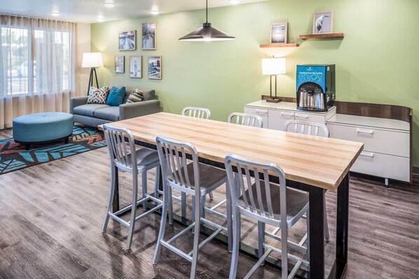 A modern hotel room with a wooden dining table, white chairs, a sofa with patterned cushions, a standing lamp, and a coffee machine. - Woodspring Suites Orlando Kissimmee Maingate (Kissimmee)