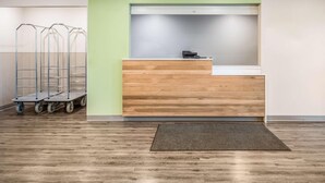 Un mostrador de recepción con detalles de madera, una pared de acento verde y una impresora sobre el mostrador. A la izquierda, se observan carros metálicos con ruedas.