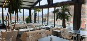 A restaurant with indoor seating, large windows, and potted plants. - Lingos Hotel (Florina)