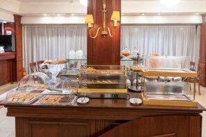 A buffet setup with various food items, a chandelier, and a dining area with a view of a curtain. - Lingos Hotel (Florina)