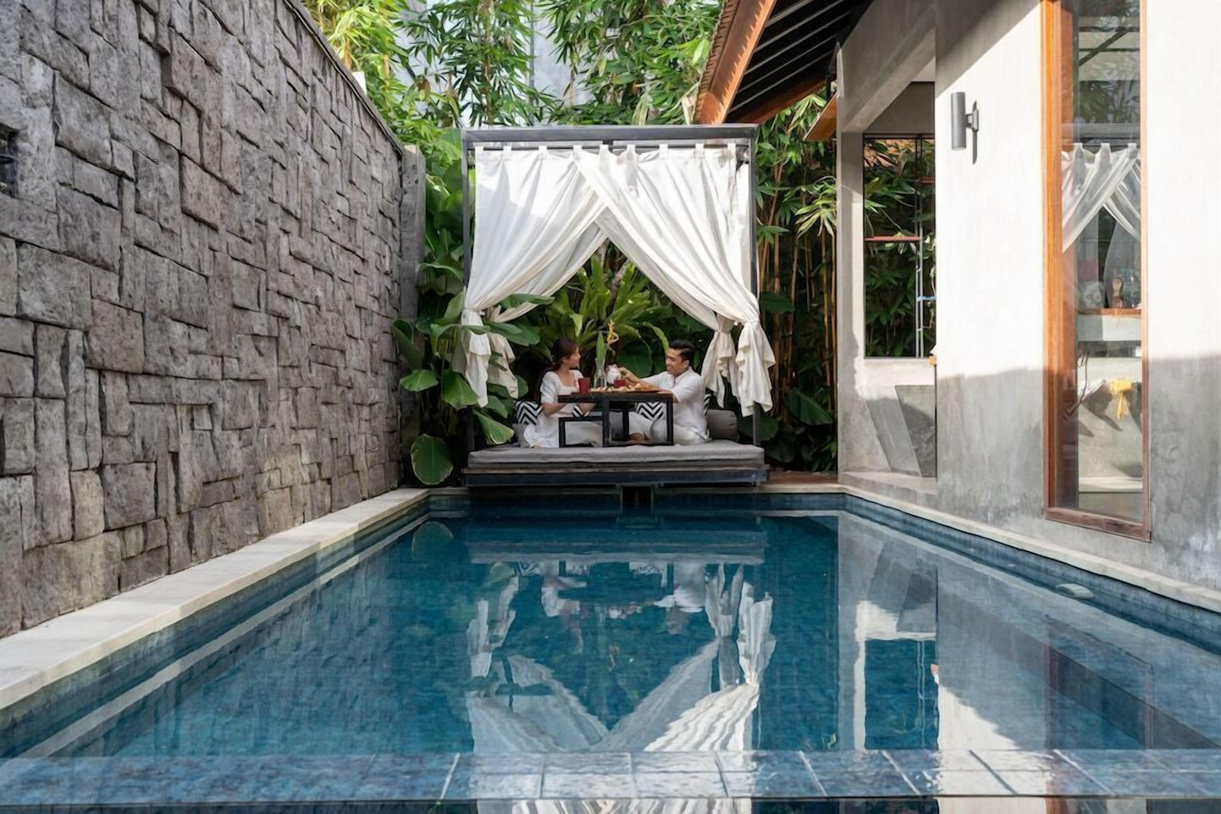 Un área de piscina con un muro de piedra, un espacio de estar cubierto y una sala con paredes de vidrio.