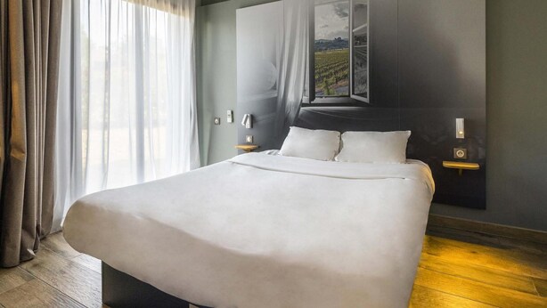 A modern hotel room with a large bed, a view of a vineyard through a window, and a dark-colored headboard. - B&B Hotel Perpignan Saleilles (Saleilles)
