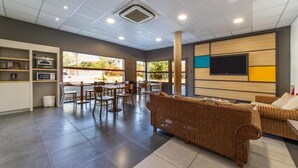 A spacious living area with a large sofa, dining table, and chairs. There is a television mounted on the wall and a kitchen area with appliances. - B&B Hotel Perpignan Saleilles (Saleilles)