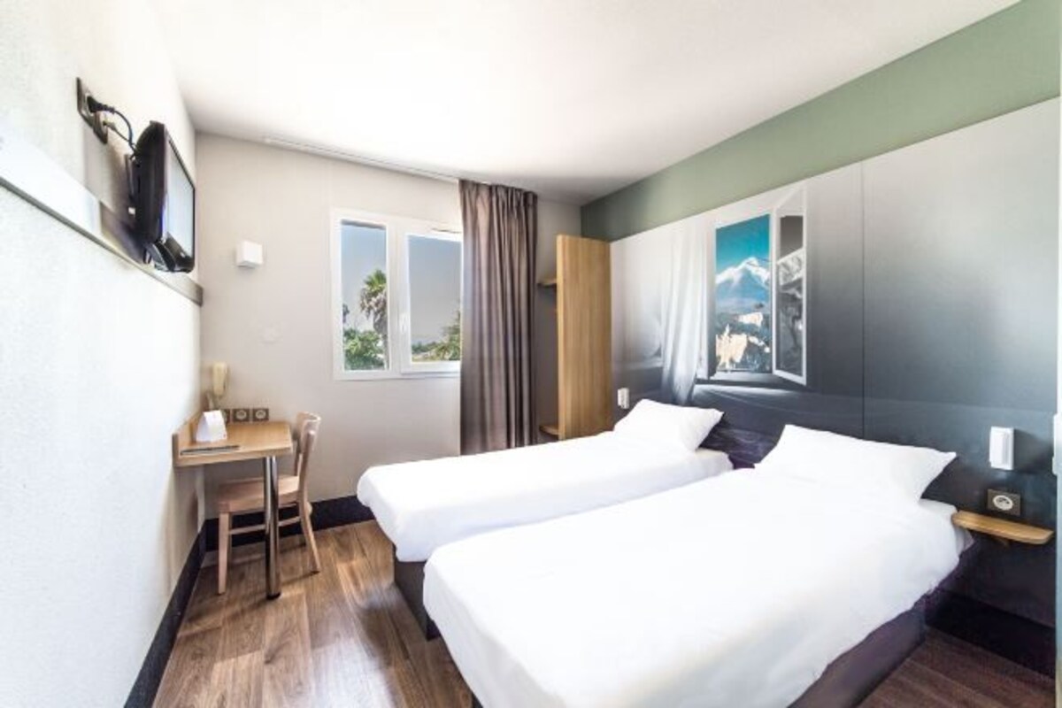 Ett hotellrum med två sängar, ett skrivbord, en stol, ett fönster med gardiner och en tavla på väggen.