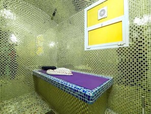 A bathroom with mosaic walls, a bathtub with a purple mat, and a small window with a yellow panel. - SHH Hotel (Al-Fujairah)