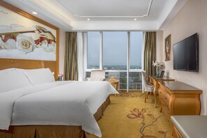 Ein Hotelzimmer mit einem großen Bett, einem Schreibtisch, einem Sessel, einem Fernseher und Blick auf die Stadt durch große Fenster.
