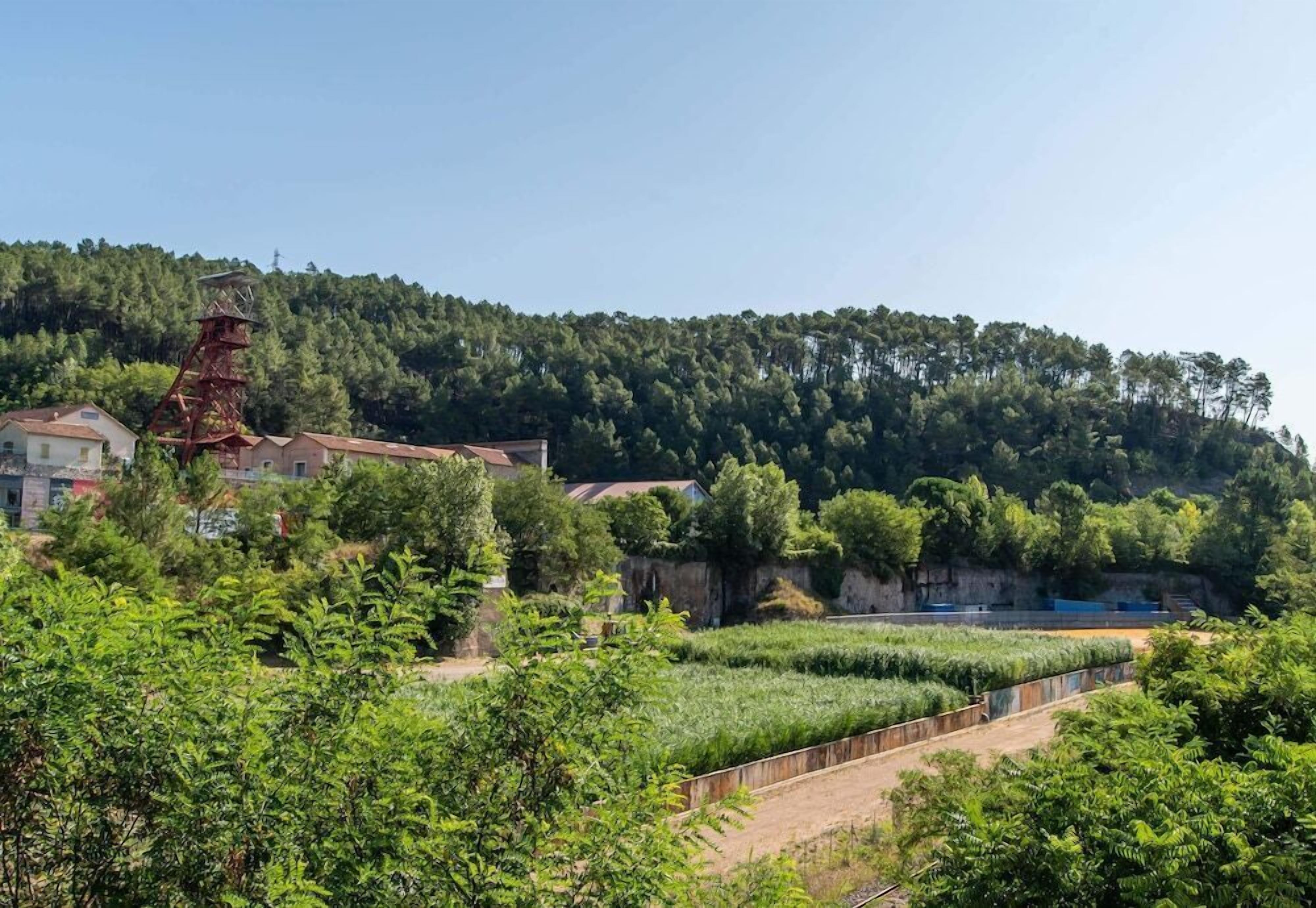 A rural landscape with a large metal structure, houses, and a forested hill.