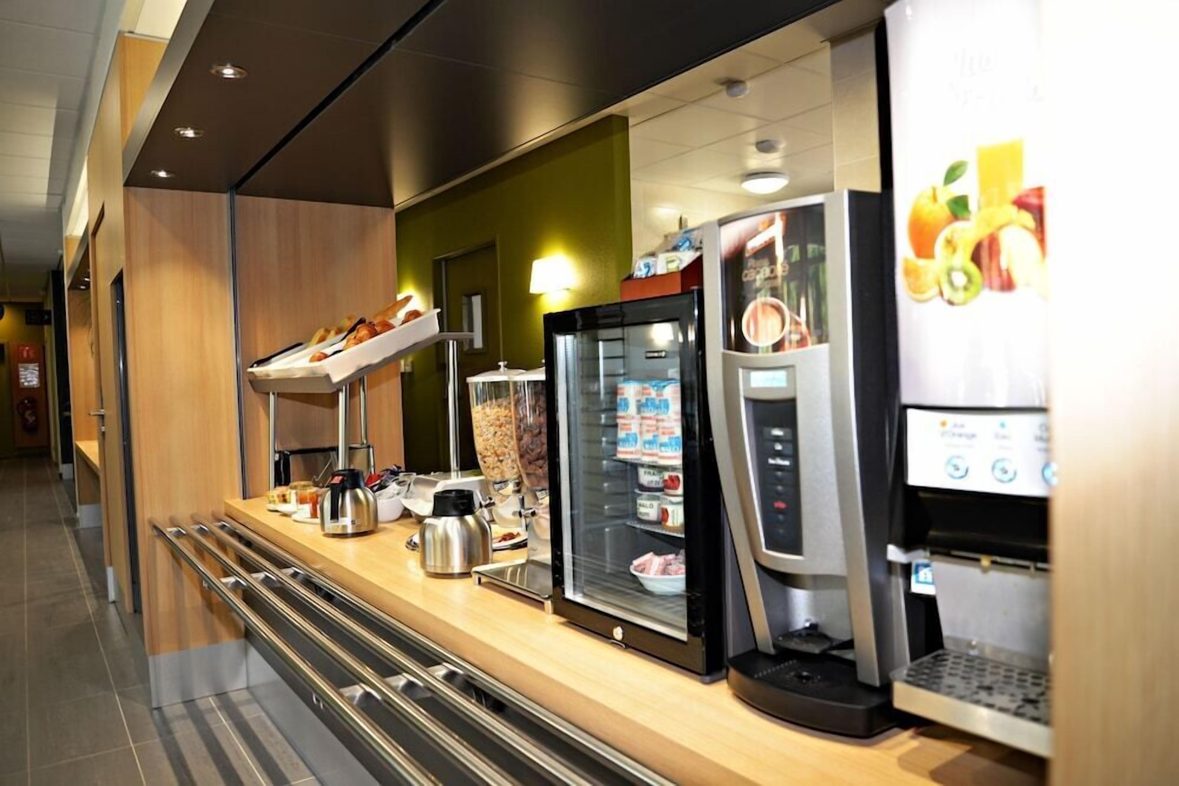 A self-service breakfast area with a coffee machine, refrigerated display case, and a beverage dispenser.