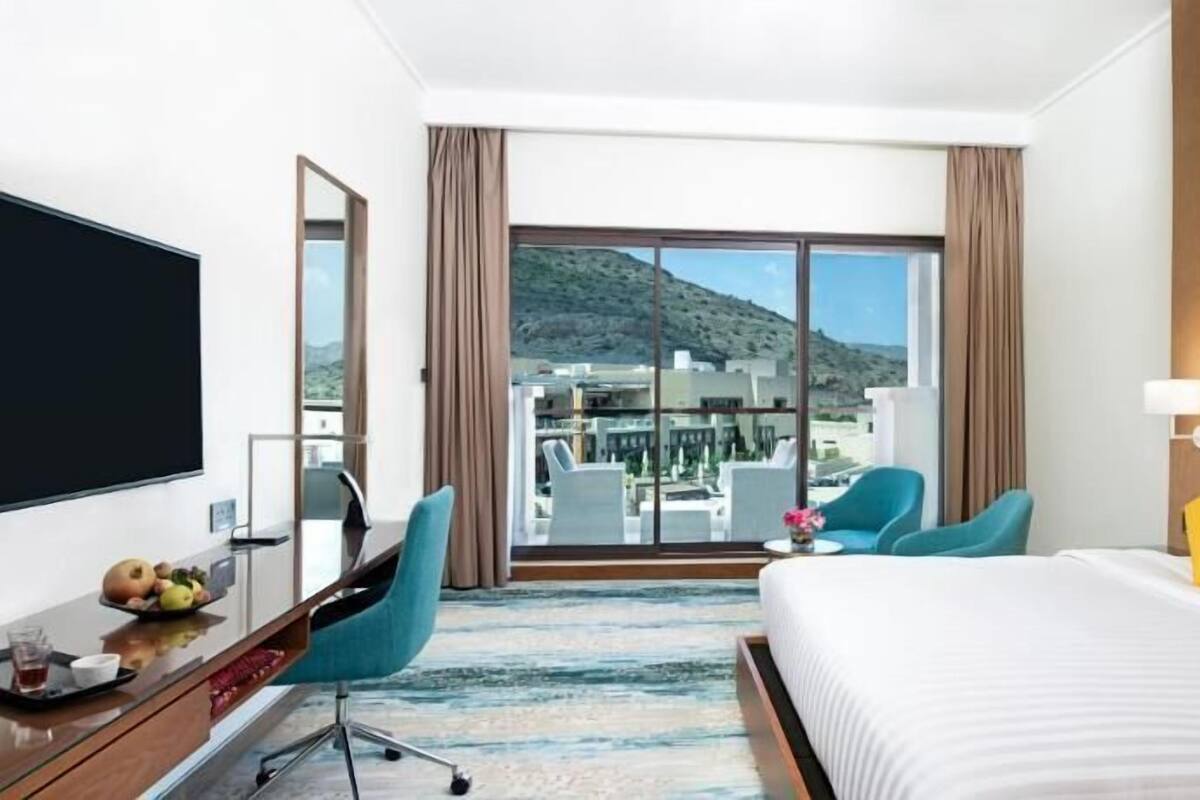 A hotel room with a bed, a desk, a chair, a TV, and a balcony with a view of buildings and hills.