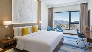 A hotel room with a large bed, a desk, two chairs, a view of mountains, and a decorative wall panel. - Dusitd2 Naseem Resort Jabal Akhdar (Sayq)