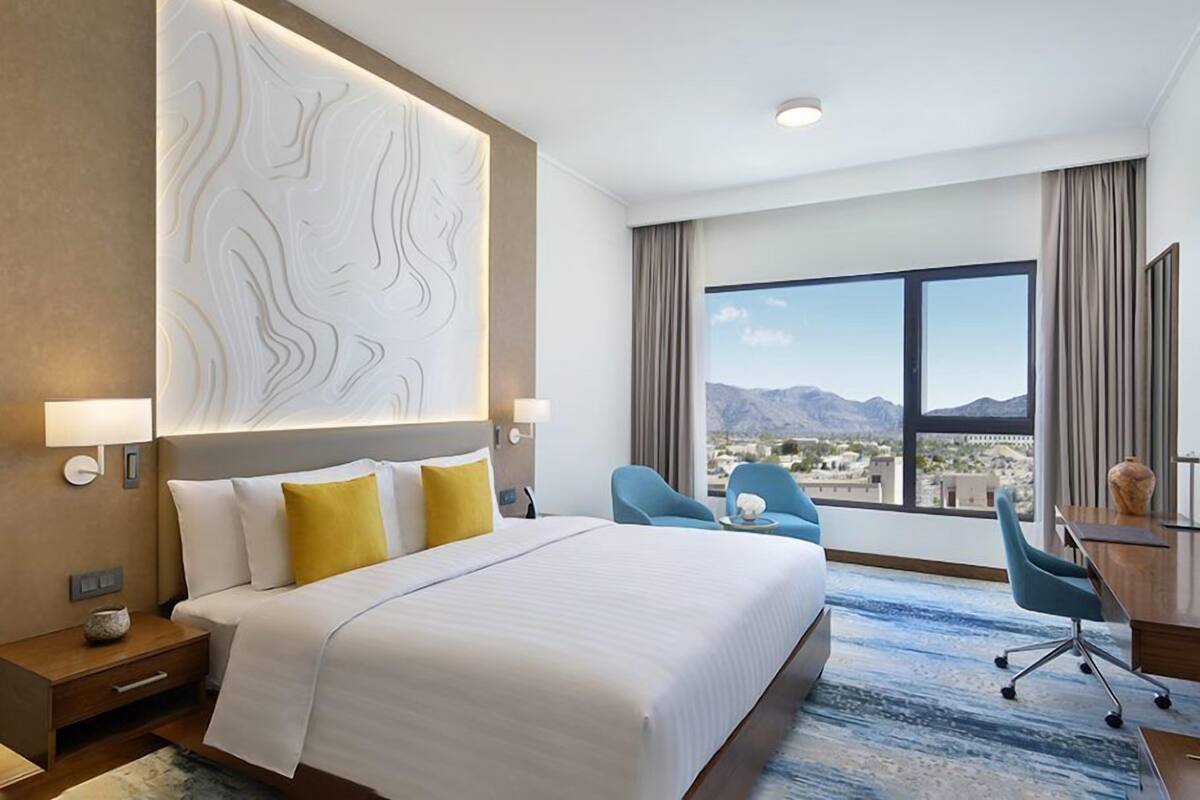 A hotel room with a large bed, a desk, two chairs, a view of mountains, and a decorative wall panel.