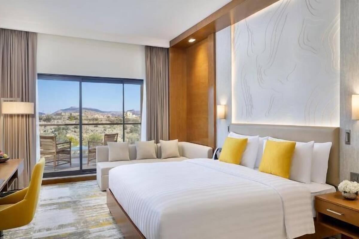 A hotel room with a large bed, a desk, a chair, and a view of a mountainous landscape.