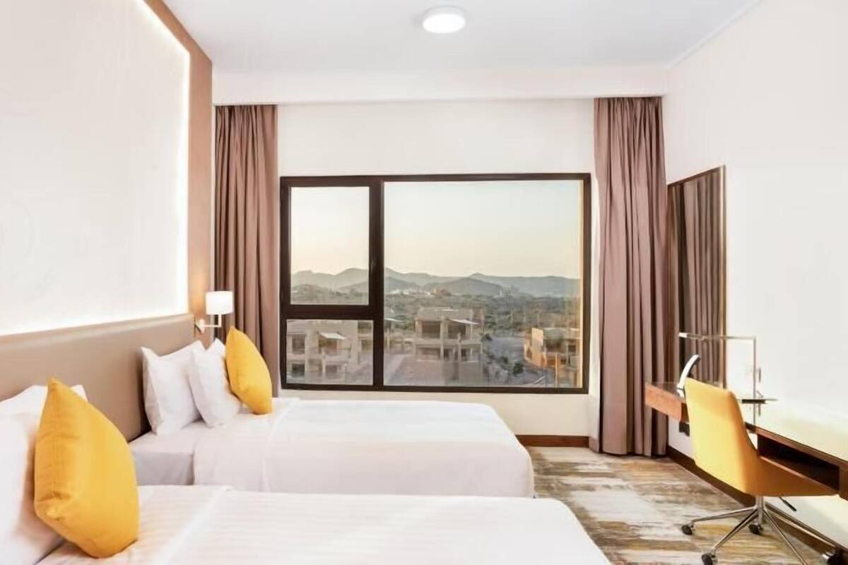 A hotel room with two beds, a desk, a chair, a large window with a city view, and curtains.