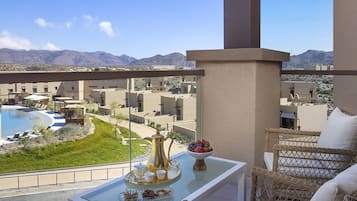 A balcony with a table set for tea, a bowl of fruit, and a view of a pool and buildings in the distance.