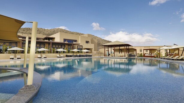 A swimming pool with lounge chairs and umbrellas, a covered seating area, and a building with a desert backdrop. - Dusitd2 Naseem Resort Jabal Akhdar (Sayq)