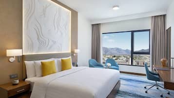 A hotel room with a large bed, a desk, two chairs, a view of mountains, and a decorative wall panel.