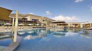 A swimming pool with lounge chairs and umbrellas, a covered seating area, and a building with a desert backdrop.
