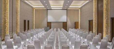 Un salón de conferencias con filas de sillas blancas, una pantalla grande y piso alfombrado.