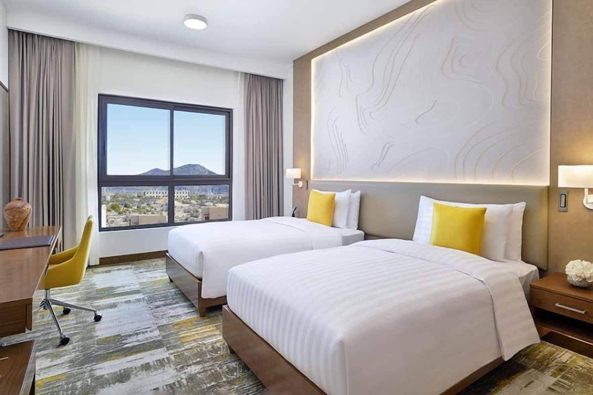 A hotel room with two beds, a desk, a chair, and a large window with a view.