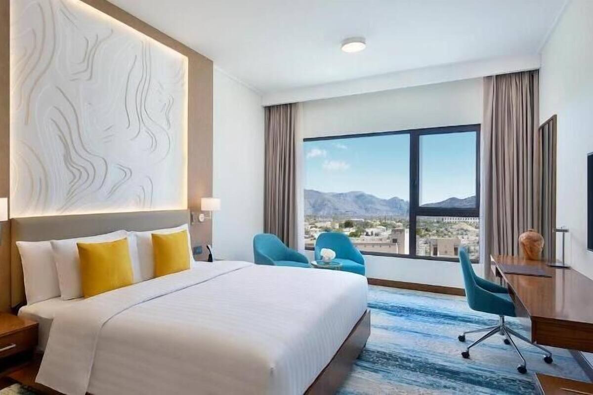 A hotel room with a large bed, two chairs, a desk, a large window with a mountain view, and a decorative wall panel.