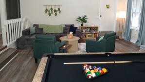 Un salon avec une table de billard, des canapés verts, une bibliothèque et une plante en pot.