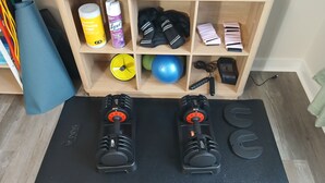 Un espace de remise en forme aménagé à la maison, comprenant deux haltères réglables, un tapis de yoga et une étagère en bois contenant du matériel et des accessoires d'exercice.