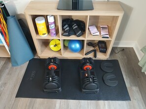 Angolo palestra in casa con due manubri regolabili, un tappetino da yoga e una mensola in legno con attrezzature e accessori per esercizi.