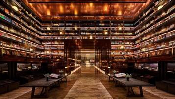 A library with bookshelves on all sides, a central aisle, and tables with plants.