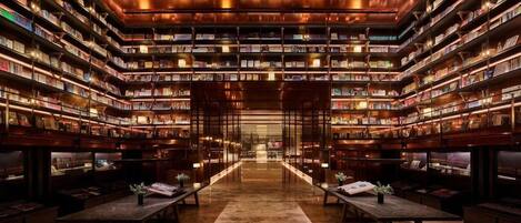 Une bibliothèque avec des étagères qui recouvrent tous les murs, une allée centrale et des tables ornées de plantes.