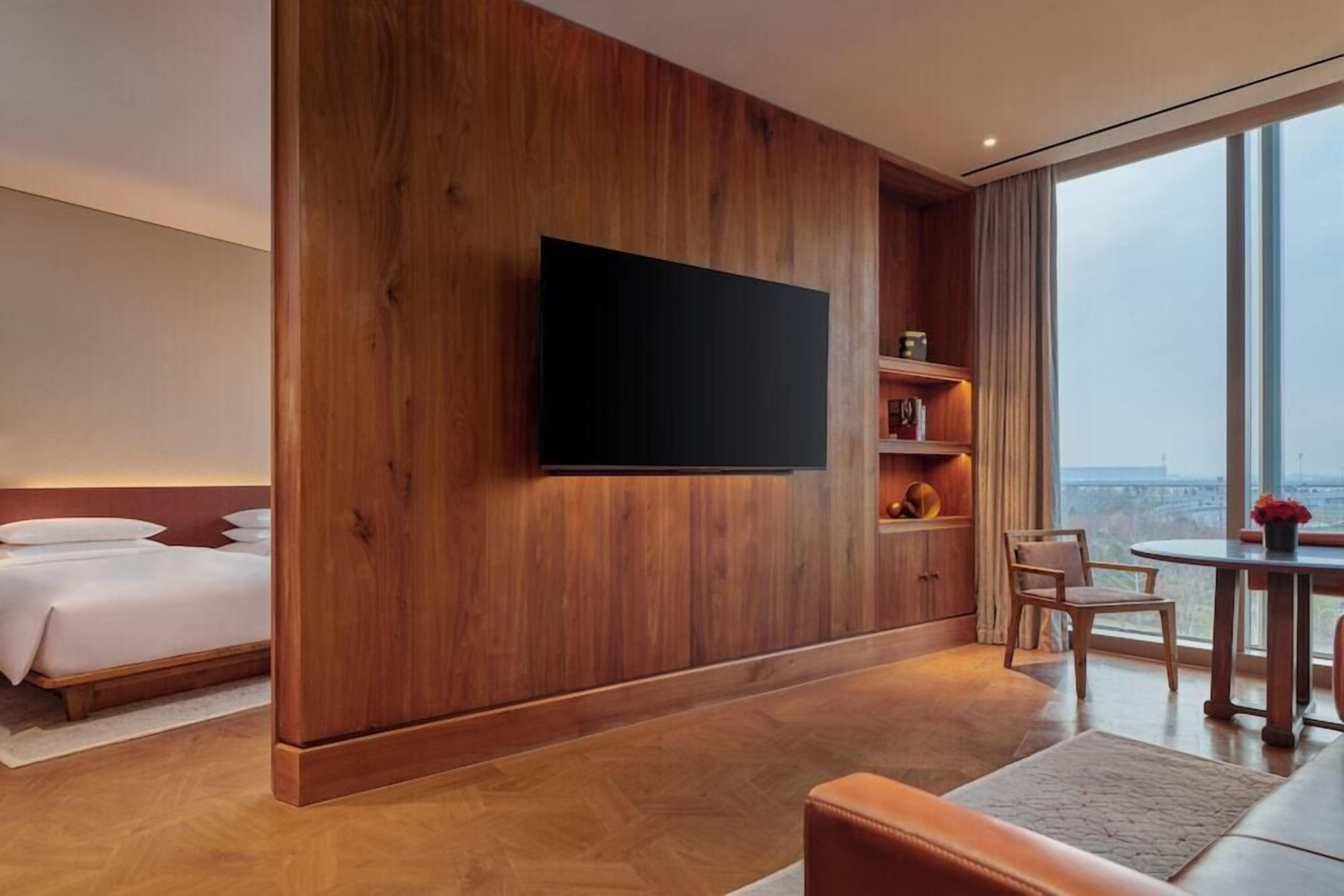 Une chambre d’hôtel moderne dotée d’un grand téléviseur fixé à un mur en bois, d’un lit avec du linge de lit blanc, d’un coin salon avec un canapé en cuir et d’une table à manger offrant une vue sur la ville grâce à de grandes fenêtres.