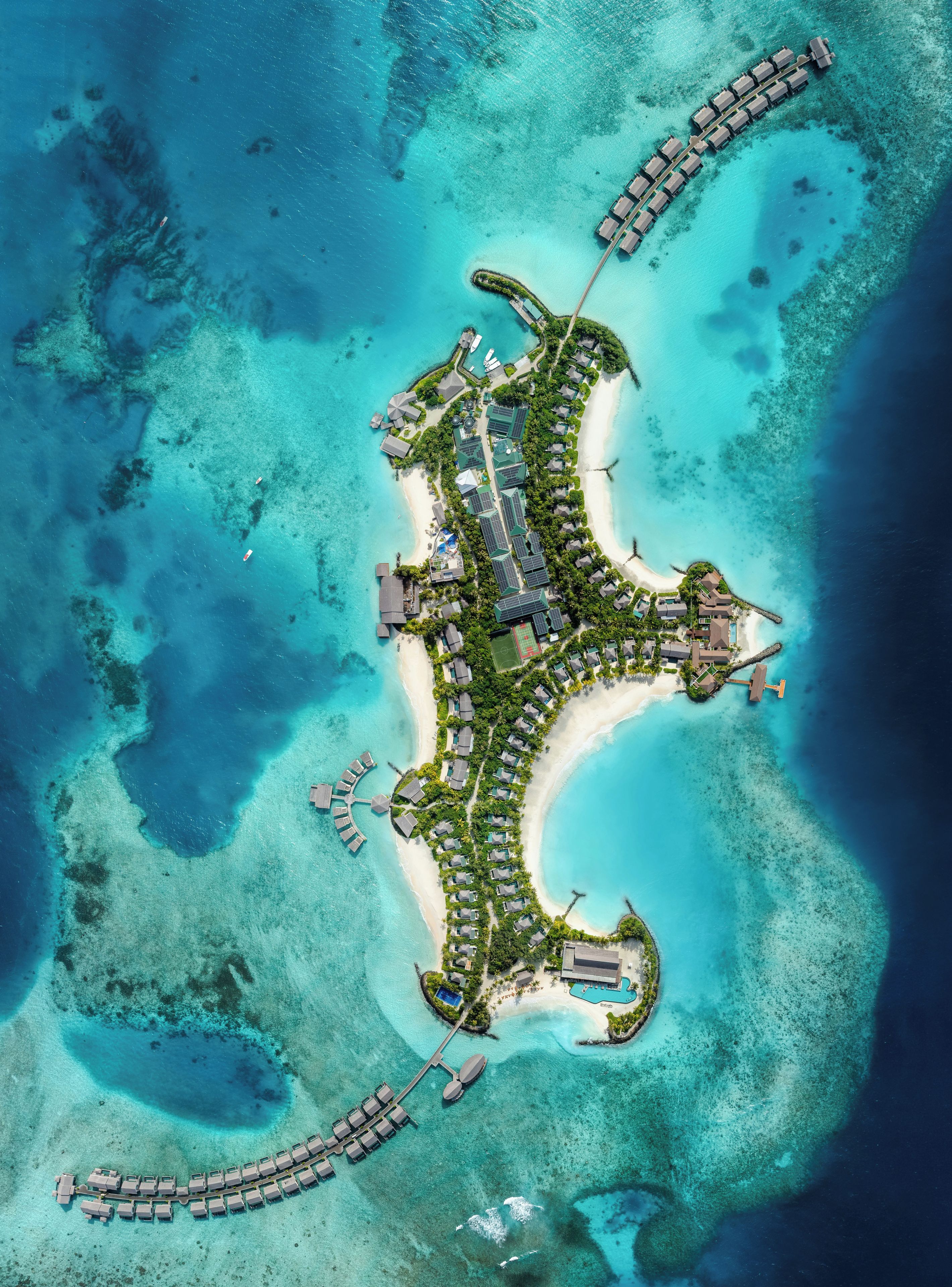 An aerial view of a resort island with a crescent-shaped layout, featuring a marina, multiple buildings, and clear turquoise waters.