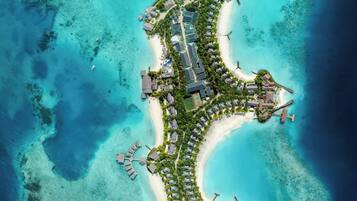 An aerial view of a resort island with a crescent-shaped layout, featuring a marina, multiple buildings, and clear turquoise waters.