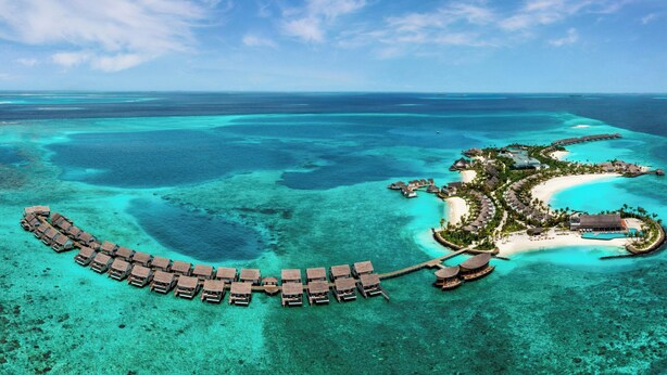 An aerial view of a resort with overwater bungalows, a beach, and clear blue waters.