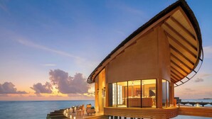 A modern overwater bungalow with a curved roof, a glass-enclosed living area, and a deck with seating.