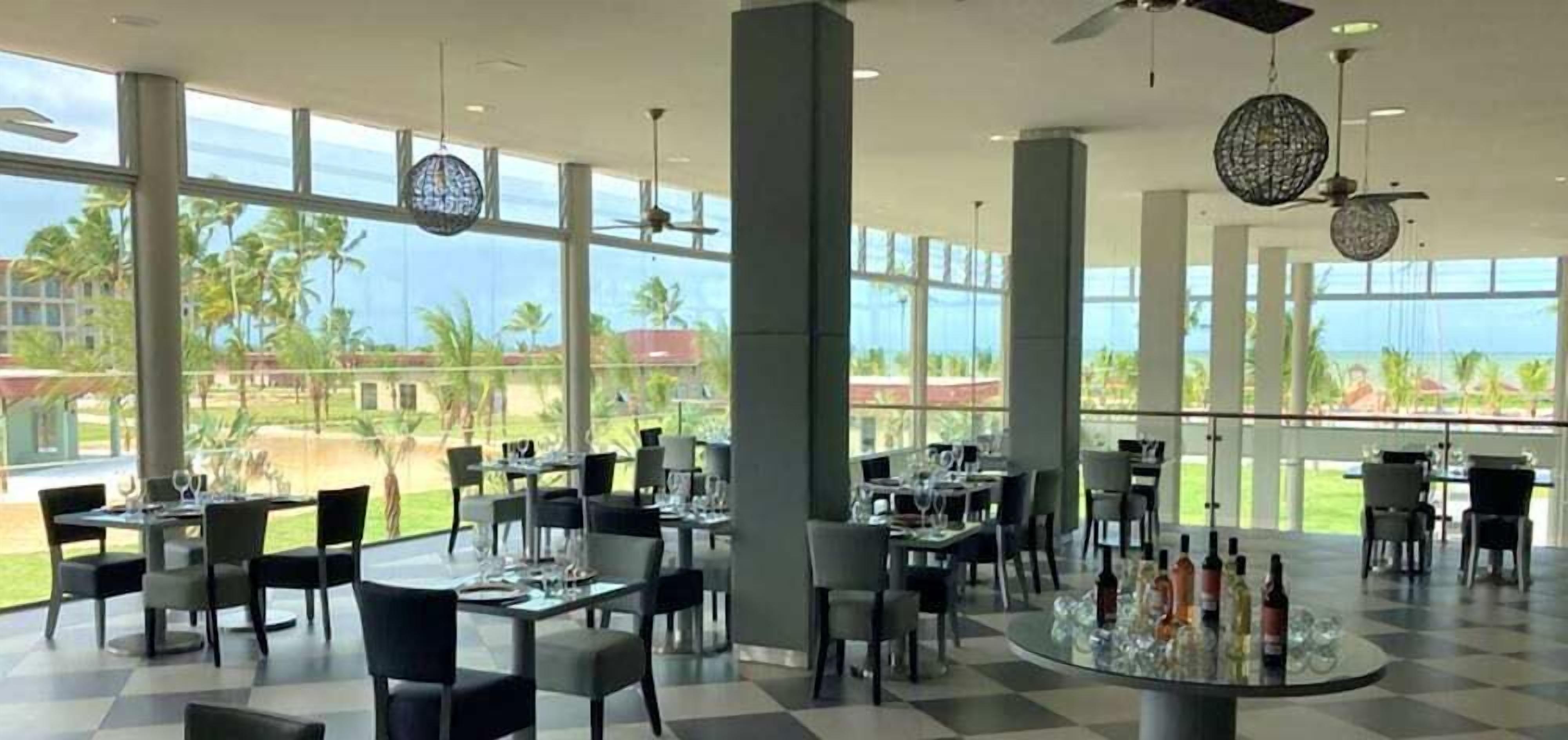 A spacious indoor dining area with large windows offering a view of palm trees and a clear sky. Tables are set with glasses and cutlery, and there are ceiling fans and decorative light fixtures.