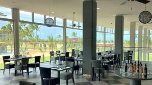 A spacious indoor dining area with large windows offering a view of palm trees and a clear sky. Tables are set with glasses and cutlery, and there are ceiling fans and decorative light fixtures. - Vila Gale Alagoas (Barra de Santo Antonio)