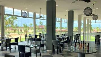 A spacious indoor dining area with large windows offering a view of palm trees and a clear sky. Tables are set with glasses and cutlery, and there are ceiling fans and decorative light fixtures.