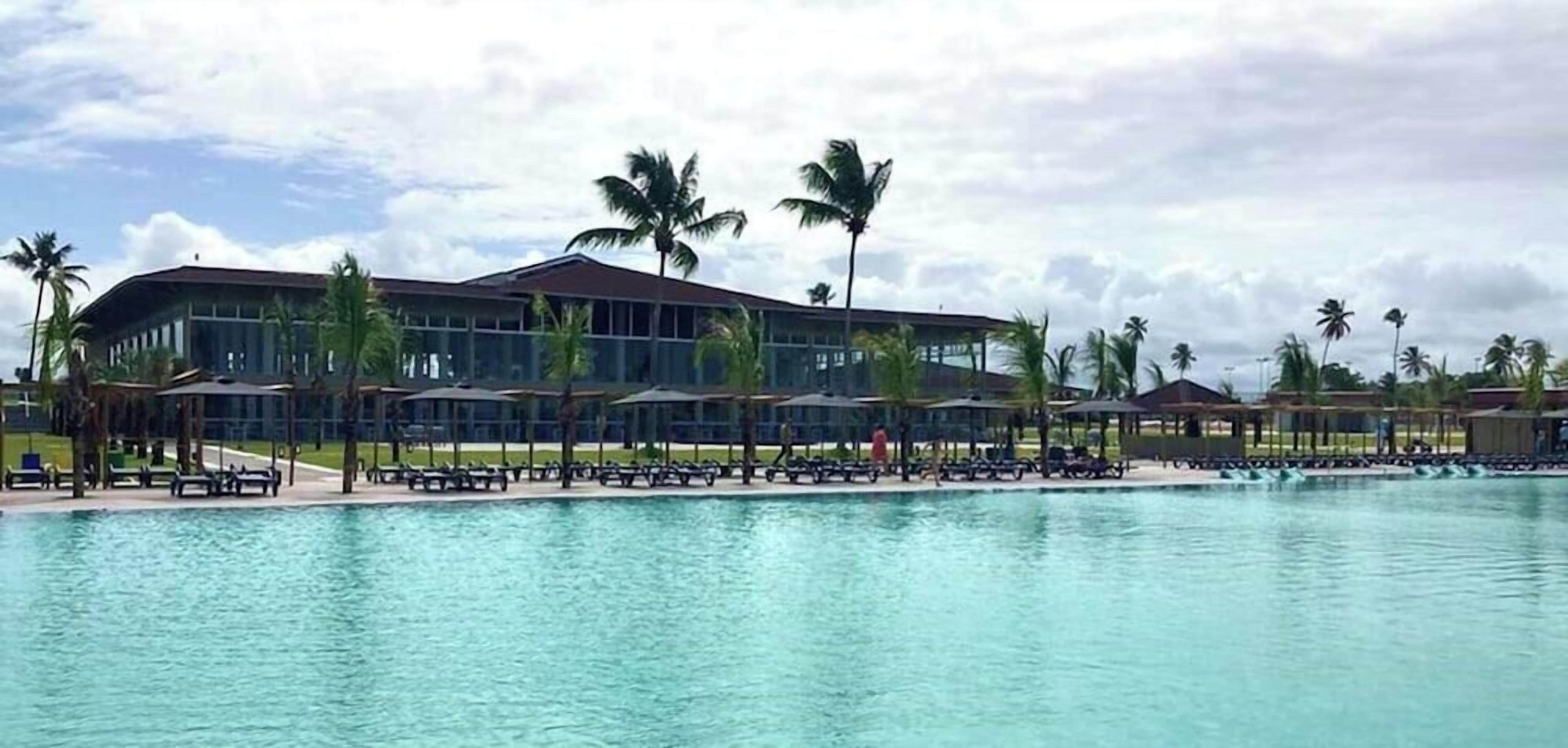 A large swimming pool with lounge chairs and umbrellas, surrounded by palm trees and a modern building.
