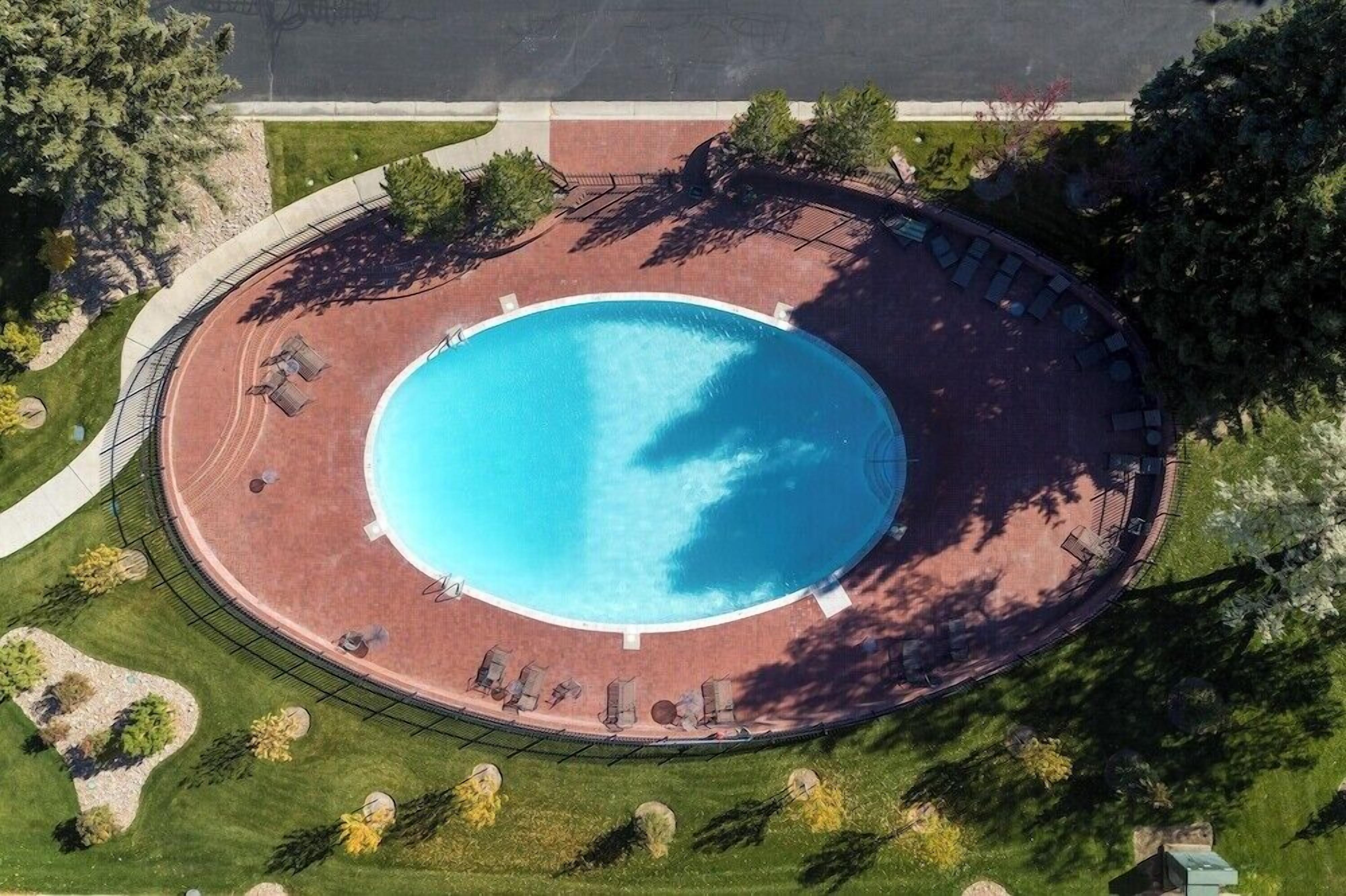An aerial view of a circular swimming pool with a surrounding deck, surrounded by trees and a paved walkway.