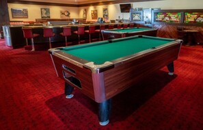 A pool table with a red carpeted floor, a bar area with red chairs, and a television screen in the background.