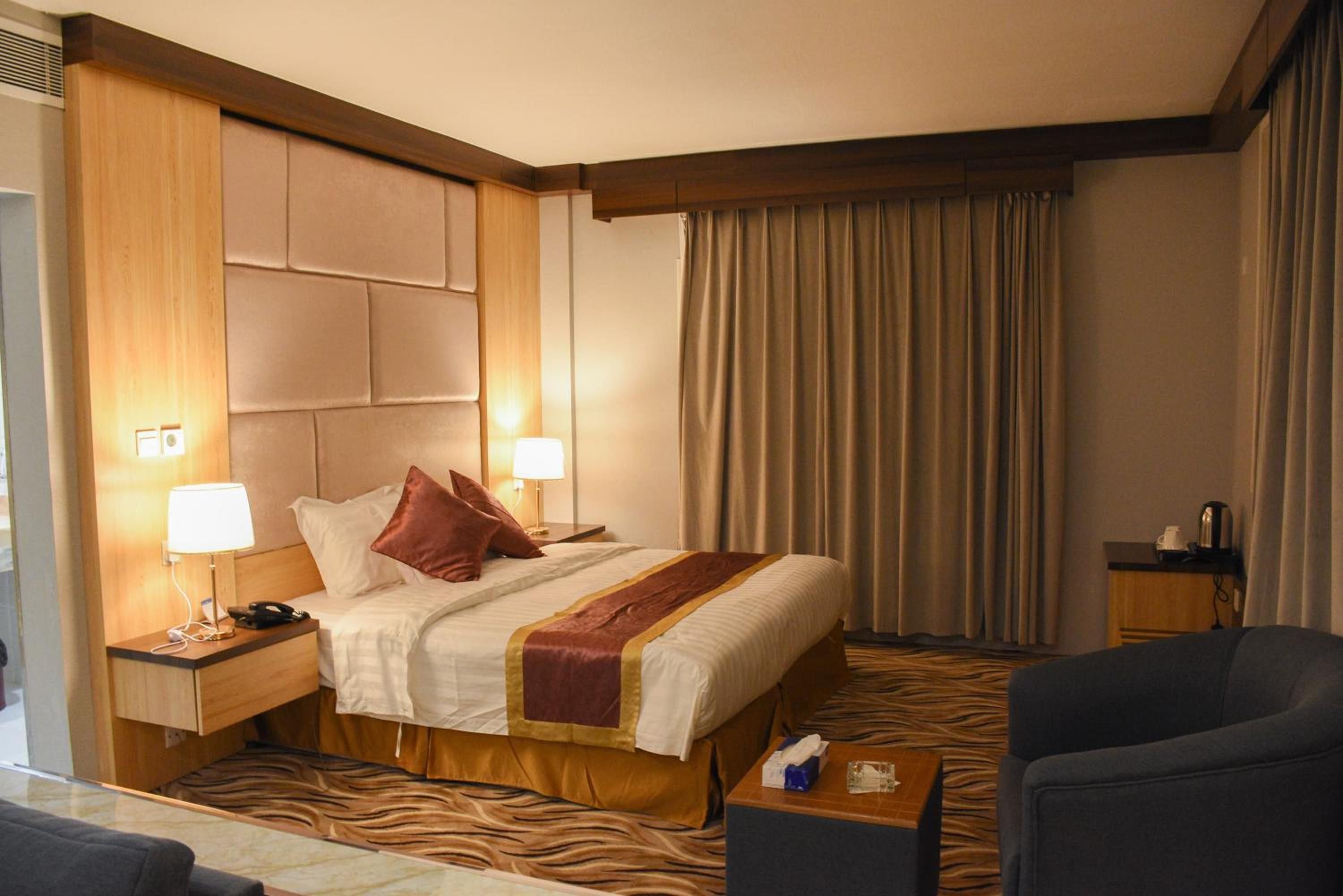 A hotel room with a bed, two bedside lamps, a nightstand, a chair, a small table, and a window with curtains.