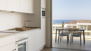 Una cocina moderna con armarios blancos, horno empotrado y microondas. Al exterior, un balcón con mesa y sillas, con vistas a un paisaje costero.