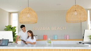 Zona de recepción con un cartel que dice "NIVARIA BEACH", dos lámparas colgantes de mimbre, un mostrador de recepción, un monitor de computadora y una planta.